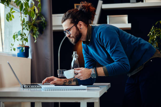 Young attractive bearded hipster with curly hair leaning on table, drinking coffee and surfing on internet while standing in home office.  curley cup stock pictures, royalty-free photos & images