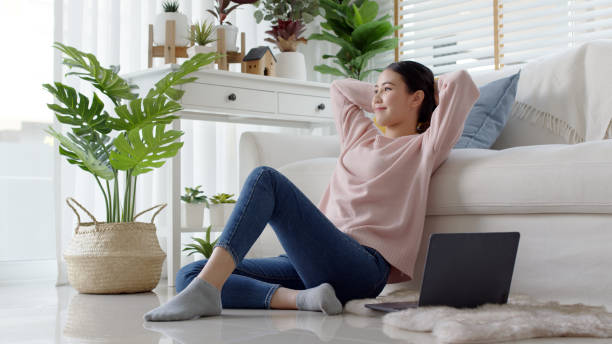 Young attractive beautiful asia female girl or university student sit smile look outside window put arm hand back behind head at sofa couch living room feeling relax comfort at cozy home houseplant. stock photo