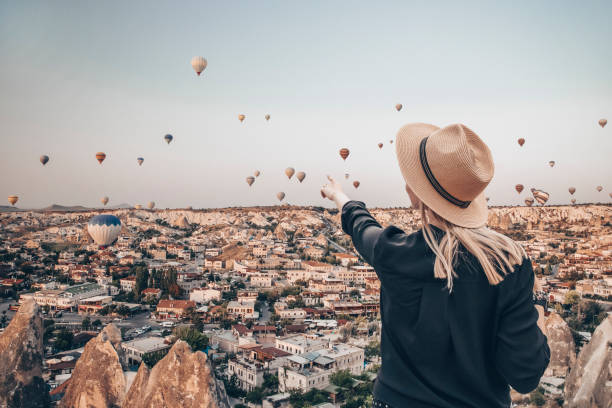 Young attractive girl in a hat stands on the mountain with flying air balloons on the background. Finger pointing girl on the sunrise. View from the back. Famous tourist Turkish region cappadocia. stock photo