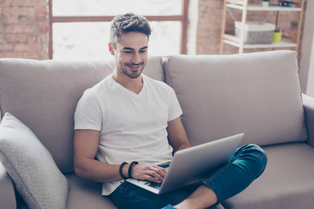 젊은 매력적인 웃는 남자 그의 노트북에서 검색, 집에서 집에서 아늑한 베이지색 소파에 앉아, 캐주얼 옷을 입고 - 청년 남자 뉴스 사진 이미지