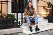 istock Young attractive woman in trench coat and jeans dreamily looking in camera sitting on little stairs spending time on cozy city street 1357102978