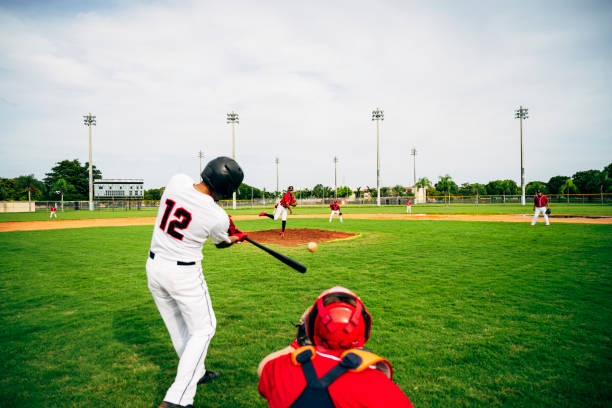 Young baseball player swinging his bat at thrown pitch Rear viewpoint of Hispanic baseball player standing in batter’s box and swinging his bat at thrown pitch. baseball ball stock pictures, royalty-free photos & images