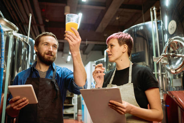 Young bearded brewer in apron looking at beer in glass while standing next to his female colleague stock photo