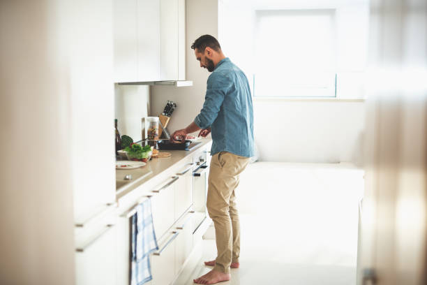 Young bearded man cooking dinner at cozy kitchen Side view full length portrait of handsome gentleman putting burger bun in frying pan barefoot photos stock pictures, royalty-free photos & images