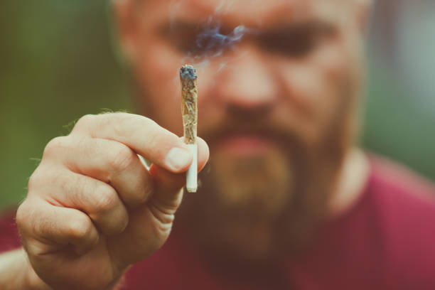 Young bearded man holding a smoking marijuana joint - legalization medical cannabis concept - smoke is coming out of the weed or hashish cigarette stock photo