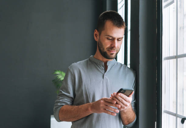 Young bearded man in gray longsleeve with smartphone in hands reading message standing at window in modern office with dark walls stock photo