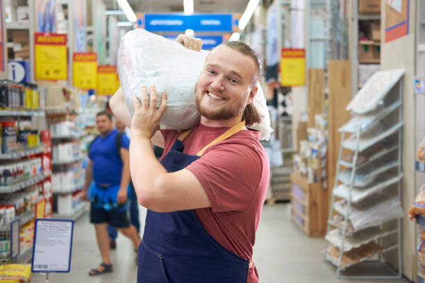 Young bearded salesman carries a pack of plaster on his shoulder in counstruction store stock photo