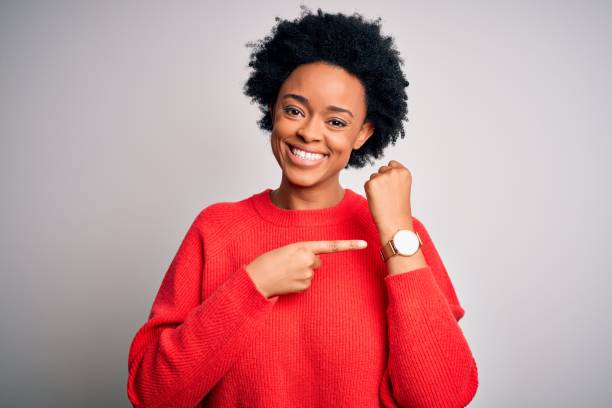 jonge mooie mooie afrikaanse amerikaanse afrovrouw met krullend haar dat rode toevallige sweater draagt in haast die aan horlogetijd, ongeduld richt, die de camera met ontspannen uitdrukking bekijkt - woman horloge stockfoto's en -beelden