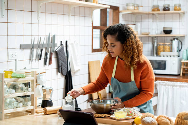 Young beautiful African American woman in kitchen learning online virtual cooking class from tablet computer at home stock photo
