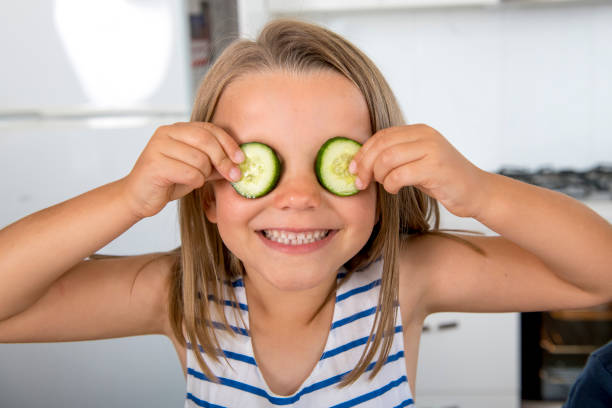young beautiful and adorable girl 6 or 7 years old having fun at home kitchen putting cucumber slices on her eyes taking tongue out happy in children lifestyle concept stock photo