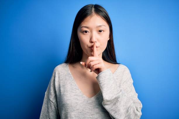 Young beautiful asian woman wearing casual sweater standing over blue isolated background asking to be quiet with finger on lips. Silence and secret concept. Young beautiful asian woman wearing casual sweater standing over blue isolated background asking to be quiet with finger on lips. Silence and secret concept. how do you say shut up in japanese stock pictures, royalty-free photos & images