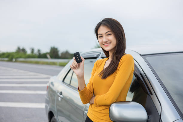 Young beautiful asian women buying new car. she was standing in near car on the roadside. Hand showing car key. Smiling female driving vehicle on the road on a bright day stock photo