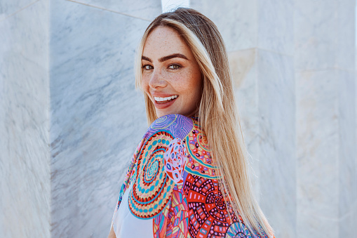 Closeup portrait of young beautiful blond woman in summertime. Female outdoors wearing colorfull smock with marble column background