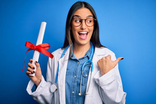 Young beautiful brunette doctor woman wearing glasses and coat holding diploma degree pointing and showing with thumb up to the side with happy face smiling Young beautiful brunette doctor woman wearing glasses and coat holding diploma degree pointing and showing with thumb up to the side with happy face smiling medical degrees stock pictures, royalty-free photos & images