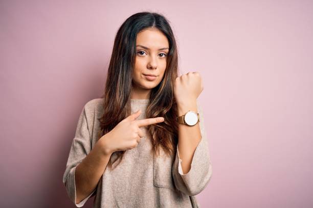 jonge mooie donkerbruine vrouw die toevallige sweater draagt die zich over roze achtergrond bevindt in haast die aan horlogetijd, ongeduld, het bekijken van de camera met ontspannen uitdrukking richt - woman horloge stockfoto's en -beelden