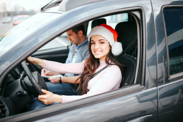 Young beautiful cheerful woman sit on driver's place in car. Holding hands on steering wheel and smile. Wear red hat. Christmas or new year period. Man sit besides her. stock photo