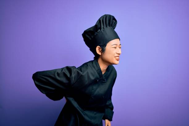 giovane bella chef cinese donna che indossa l'uniforme del fornello e cappello su sfondo viola sofferenza di mal di schiena, toccare con la mano, dolore muscolare - chef backache foto e immagini stock