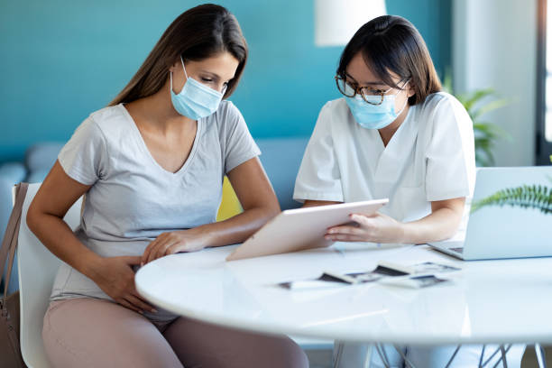 jonge mooie gynaecoloog die een hygiënisch gezichtsmasker draagt terwijl het tonen aan zwangere vrouw ultrasone scanbaby met digitale tablet in medisch overleg toont. - arts vrouw mondkapje stockfoto's en -beelden