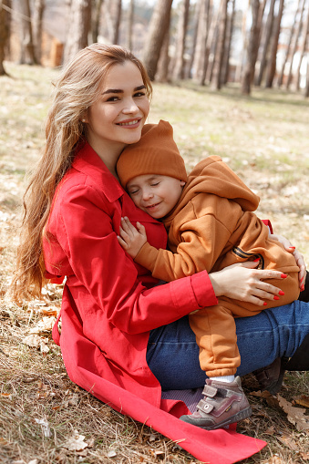 Happy young beautiful mother sitting on the ground in the park and holding his son. Little happy child boy snuggles up to his beloved mom. Sunny family portrait in the forest, spring or autumn