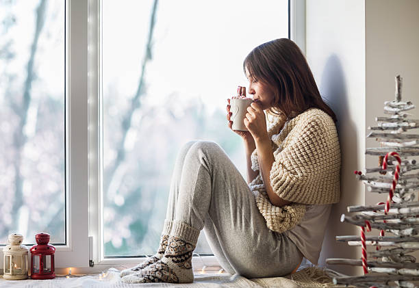 Young beautiful woman drinking hot coffee sitting on window sill Young beautiful woman drinking hot coffee sitting on window sill in christmas decorated home. Holiday concept sunday morning coffee stock pictures, royalty-free photos & images