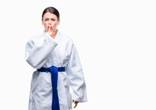 Young beautiful woman wearing karate kimono uniform over isolated background asking to be quiet with finger on lips. Silence and secret concept. Young beautiful woman wearing karate kimono uniform over isolated background asking to be quiet with finger on lips. Silence and secret concept. how do you say shut up in japanese stock pictures, royalty-free photos & images