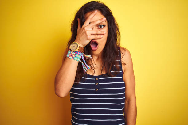 Young beautiful woman wearing striped t-shirt standing over isolated yellow background peeking in shock covering face and eyes with hand, looking through fingers with embarrassed expression. Young beautiful woman wearing striped t-shirt standing over isolated yellow background peeking in shock covering face and eyes with hand, looking through fingers with embarrassed expression. shock stock pictures, royalty-free photos & images