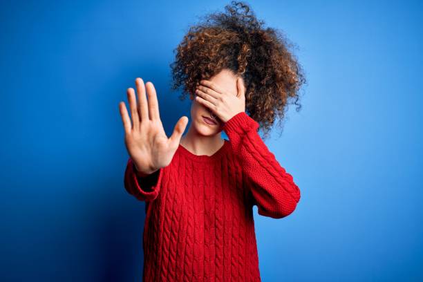 Young beautiful woman with curly hair and piercing wearing casual red sweater covering eyes with hands and doing stop gesture with sad and fear expression. Embarrassed and negative concept. stock photo