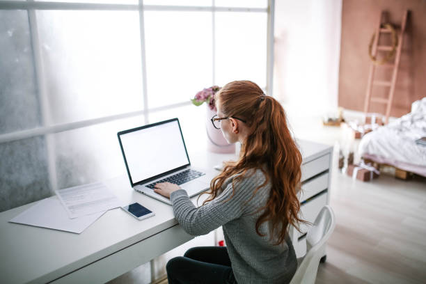 young beautiful woman with red hair, wearing glasses, working in the office, uses a laptop and mobile phone - mobile phone imagens e fotografias de stock