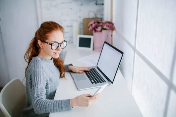 young beautiful woman with red hair, wearing glasses, working in the office, uses a laptop and mobile phone - mobile phone imagens e fotografias de stock