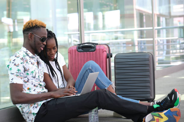 Young black couple checking their travel schedule on a laptop, sitting at the station with their suitcases. Vacation concept. stock photo