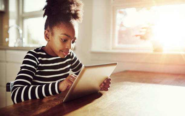Young black girl browsing on a tablet-pc Young black girl with a fun afro hairstyle sitting at a table at home browsing the internet on a tablet computer with bright sun flare through the window alongside her surfing the net stock pictures, royalty-free photos & images