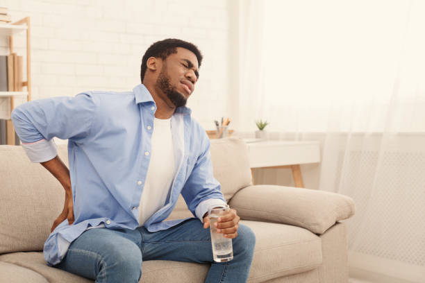 Young black man with back pain at home Young african-american man with back pain, pressing on hip with painful expression, sitting on sofa at home with glass of water, copy space back pain stock pictures, royalty-free photos & images