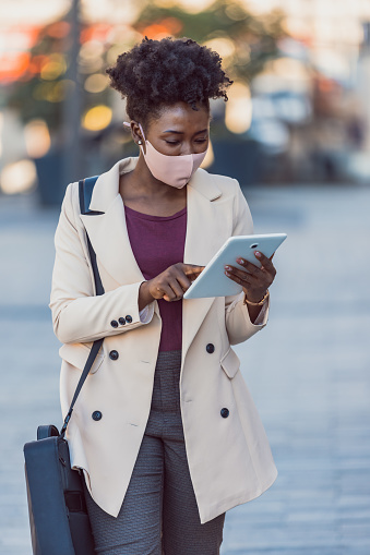 Young attractive elegant businesswoman with protective mask using touchpad while standing against modern office building.