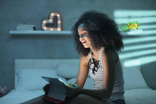 Young Black Woman Watching Movie With Laptop At Night Happy african american teenager in bed at home with tablet. Young black woman watching movie in bedroom at night and laughing. electrical equipment photos stock pictures, royalty-free photos & images