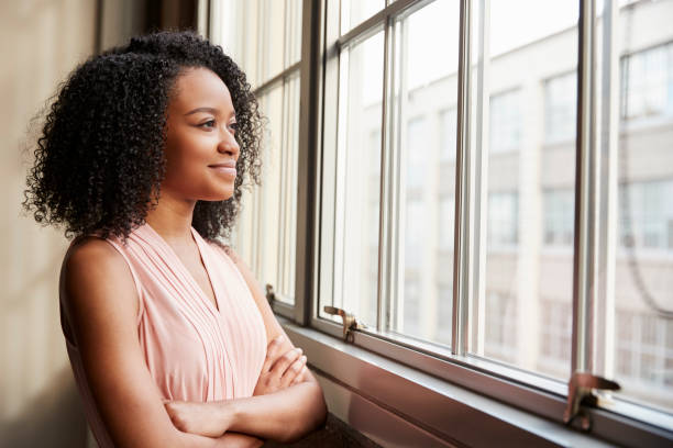 giovane donna di colore con le braccia incrociate guardando fuori dalla finestra - guardare fuori dalla finestra foto e immagini stock