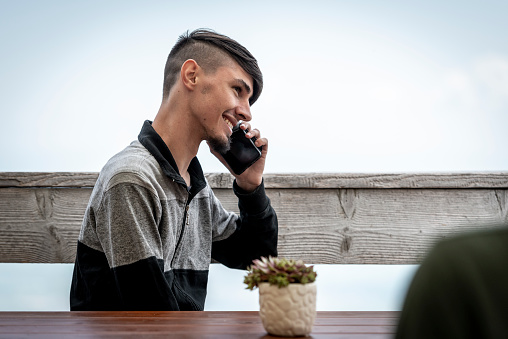 A young black-haired boy in a black-gray jacket is happy to sit at a table and make a phone call with his mobile phone and text each other.