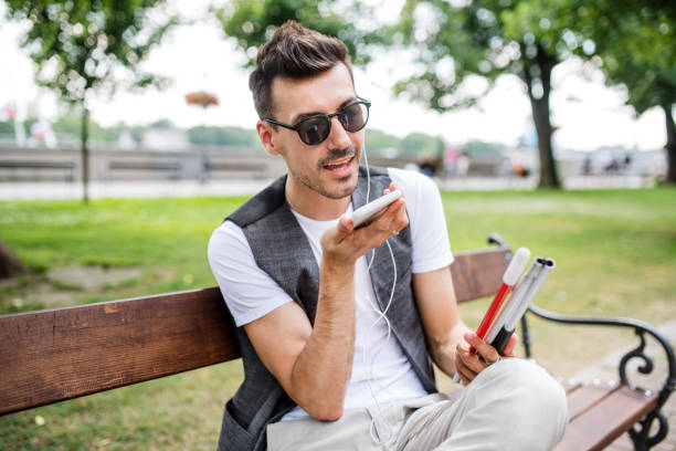 joven ciego con teléfono inteligente sentado en el banco en el parque de la ciudad, llamando. - ceguera fotografías e imágenes de stock