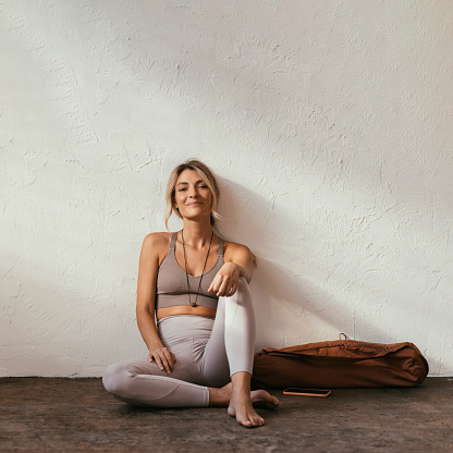 A portrait of a young blonde Caucasian woman sitting and relaxing after a yoga training looking at camera and smiling with her sports bag and her smartphone