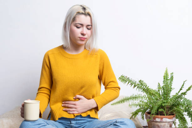 Young blonde female sitting at home holding a cup of tea while putting her hand in her stomach stock photo