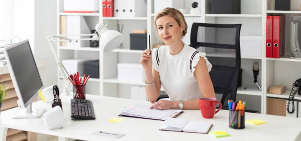 A young blonde girl is sitting at a computer desk in the office, holding a pencil in her hand and working with documents. Beautiful young girl with blond hair in a white shirt is working in the office. photo with depth of field choosing an lawyer stock pictures, royalty-free photos & images