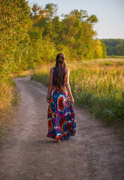 Young boho (hippie) girl is going away by dirt road in field. Rear view. Outdoor portrait at sunset time stock photo