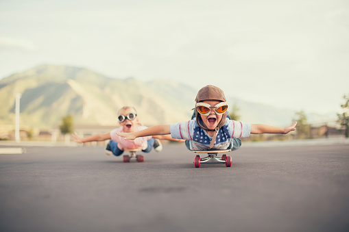 A young boy and girl are wearing flying goggles while outstretching their arms to attempt flying while on skateboards. They have large smiles and are imagining taking off into the sky.