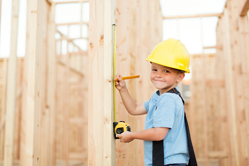 A young boy dreams of being a construction worker and building things. He is wearing a hard hat, suspenders and a tool belt while measuring on a stud of a house being built in Utah, USA.
