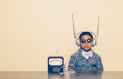 A young boy imagines reading mind and receiving communications from outer space with a homemade science project on his head. He is dressed in casual clothing, glasses and bow tie. He is wearing headphones in front of a beige background with a serious expression waiting. Retro styling.