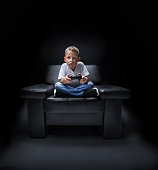 young boy in armchair watching TV and is playing with controller in his hand in front of black background