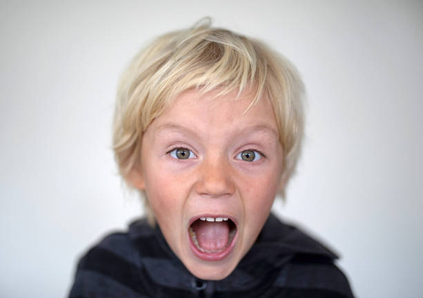 A young boy pose in front of a white screen. He is shouting at the camera with his mouth wide open stock photo