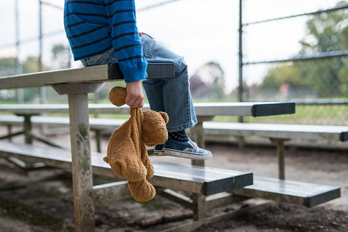 Young boy sitting by himself on bleachers and holding teddy bear.