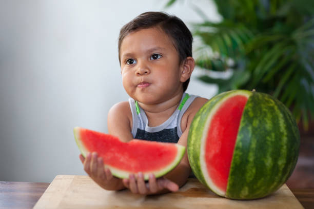 A young boy sitting in a patio, has taken a bite out of a slice of juicy seedless watermelon. stock photo
