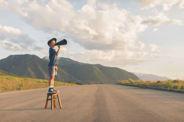 Young Boy Speaks through Megaphone A young boy dressed in paper boy hat and retro attire stands on a stool calling out his message through a megaphone. He is on a rural road in the mountains of Utah County, Utah, USA. persuasion stock pictures, royalty-free photos & images