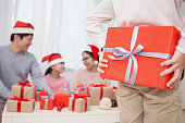 若い男の子は、彼の背中の後ろに赤いプレゼントボックスとクリスマスのための贈り物で彼の家族を驚かせる